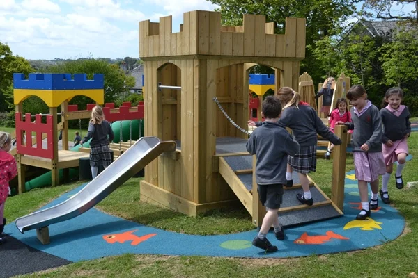 A group of children in school uniform playing on a wooden Mini Fort castle play unit with battlements, a stainless steel slide, and a drawbridge staircase, on a colourful rubber safety surface with fish motifs.