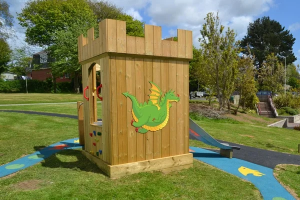 A wooden Mini Fort castle play unit with battlements, a colourful green dragon panel on the side, and a stainless steel slide, installed on a grass playground with a colourful rubber surface surround.