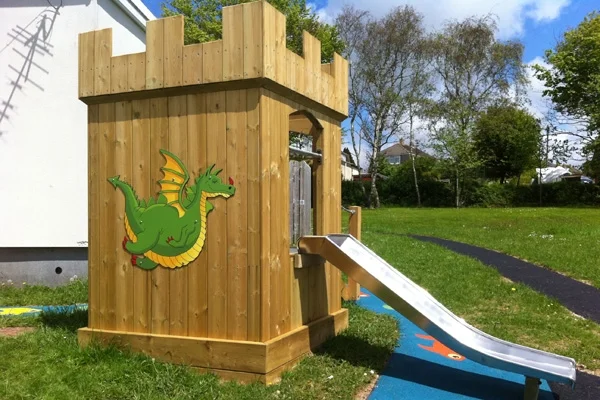 A wooden Mini Fort castle play unit with battlements, a colourful green dragon panel on the side, and a stainless steel slide, installed on a grass playground with a colourful rubber surface surround.