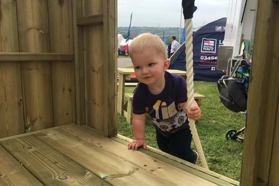 A smiling toddler in a navy t-shirt climbing a thick rope inside the elevated platform of a wooden Mini Fort castle play unit, at an outdoor show or event.