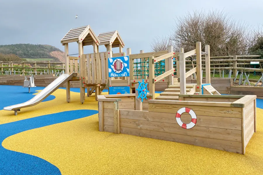 The Mini Harbour Towers coastal playground installation showing the twin-tower play unit with slide, rope net, ship's wheel, and a wooden boat-shaped play unit with lifebuoy, on a yellow and blue wave-patterned rubber surface.