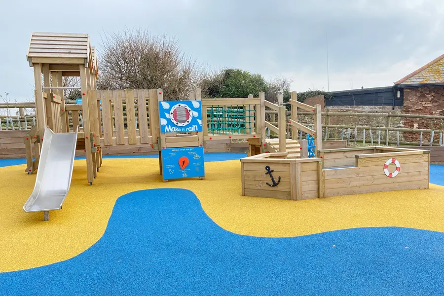A wide-angle view of the Mini Harbour Towers coastal playground showing the play tower with slide, interactive panels, rope net, and a wooden boat-shaped sandpit with anchor and lifebuoy, on a yellow and blue rubber surface.