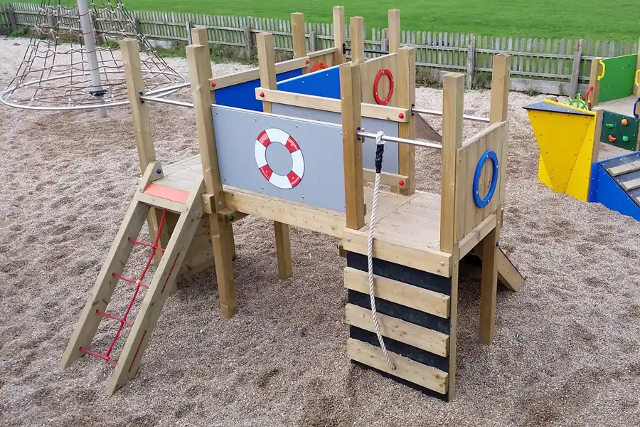 The Mini Harbour Towers nautical play unit on a sand playground, showing the elevated deck with lifebuoy panel, blue side panels, porthole rings, a rope ladder, and a red rope climbing ramp.