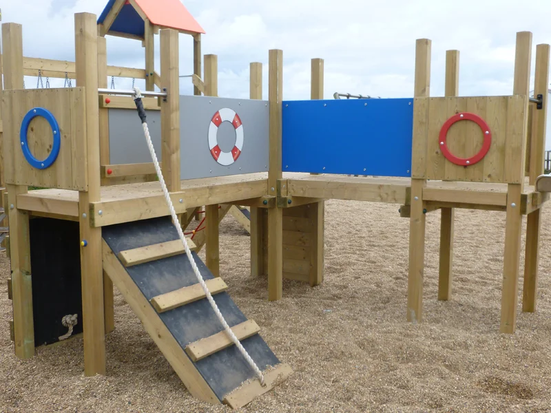 The Mini Harbour Towers nautical-themed play unit showing the elevated deck with lifebuoy panel, blue and grey side panels, porthole rings, and a rope-assisted ramp, installed on a sand playground.