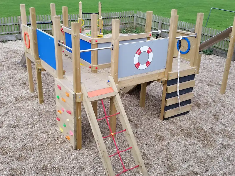 An aerial view of the Mini Harbour Towers showing the elevated deck with lifebuoy panel, blue and grey side panels, porthole rings, a colourful rock climbing wall, and a red rope ladder, on a sand playground.