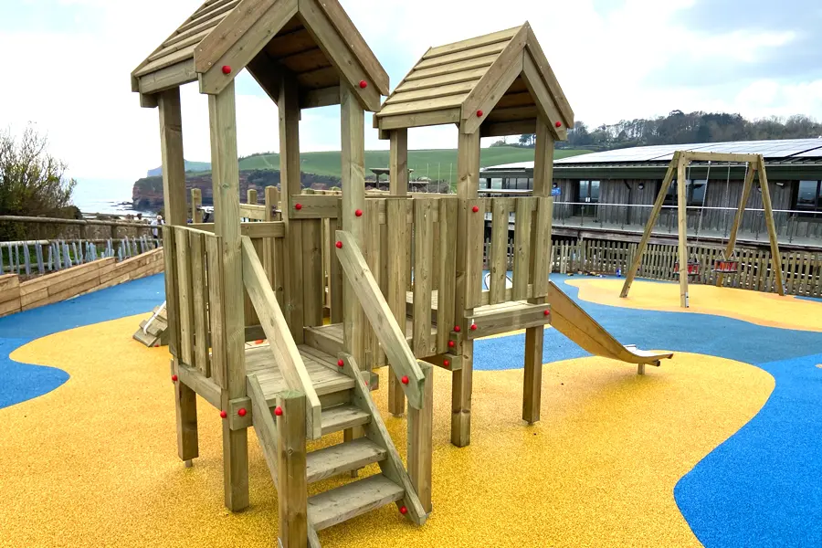 The Mini Harbour Towers twin-tower play unit with two pitched-roof towers, a central staircase with red bolt caps, and a stainless steel slide, installed on a yellow and blue wave-patterned rubber surface at a coastal playground.