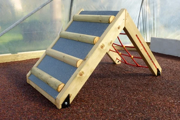 The ramp side of the Mini Ramp Net Climber, featuring a grey non-slip surface and wooden steps, on a red rubber crumb surface.