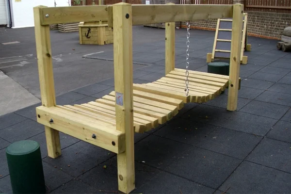 A wooden Mini Wobbly Bridge with a single central chain, installed on a black rubber tile surface in a school playground.