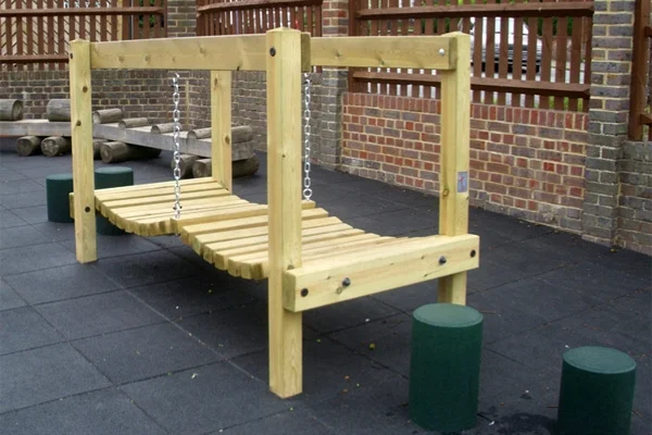 A side view of the wooden Mini Wobbly Bridge with two chains, highlighting its construction and the surrounding brick wall.