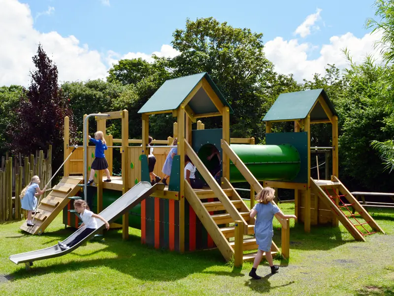 A group of children in school uniform playing on the Nature Towers multi-play unit, with a child sliding down the stainless steel slide, others climbing, and the green-roofed towers and cylindrical tunnel visible.
