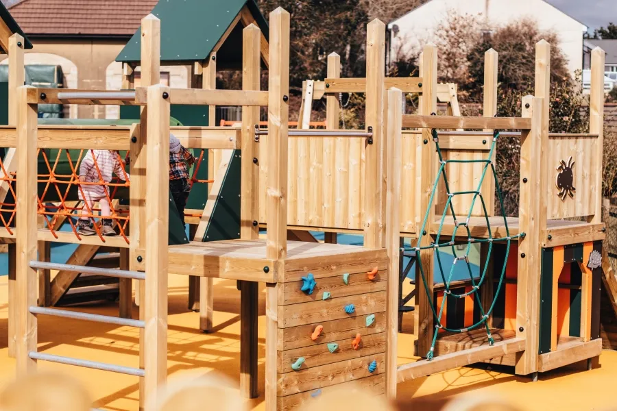 A close-up of the Nature Towers multi-play unit showing the colourful rock climbing wall with multi-coloured holds, teal rope net, galvanised ladder, and timber frame, on a yellow rubber safety surface.