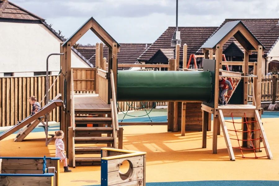 The Nature Towers multi-play unit on a grass playground, showing the colourful rock climbing wall, teal rope net, green cylindrical tunnel, monkey bars, and timber frame with green roofed towers.