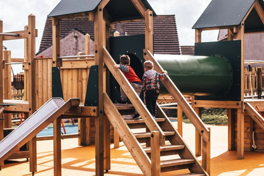Two boys climbing the wooden stairs of the Nature Towers multi-play unit towards the green cylindrical tunnel connecting two dark-roofed towers, on a yellow rubber safety surface.