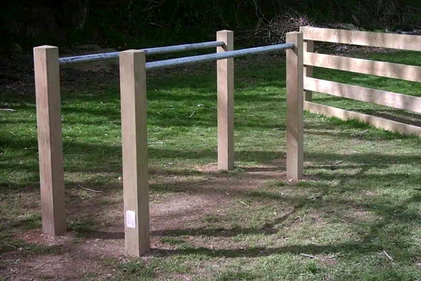A set of wooden and metal Parallel Bars outdoor play equipment next to a wooden fence in a grassy playground.