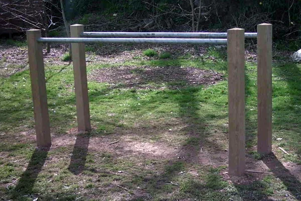 A set of wooden and metal Parallel Bars outdoor play equipment installed in a grassy park area with dappled sunlight.