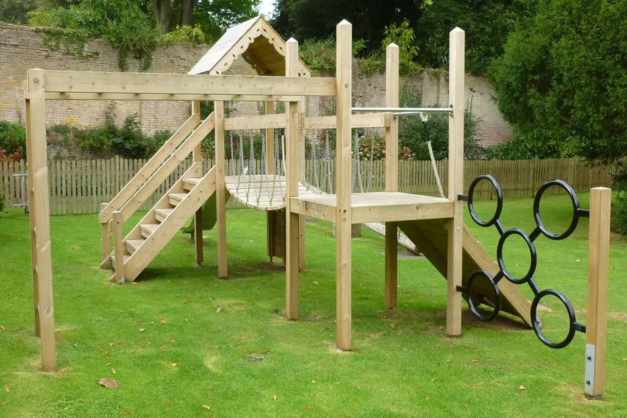 A rear view of the Pencarrow Towers multi-play unit showing the elevated platform, chain rope net, staircase access, black ring climber, and pitched-roof tower, in a walled garden with a picket fence.
