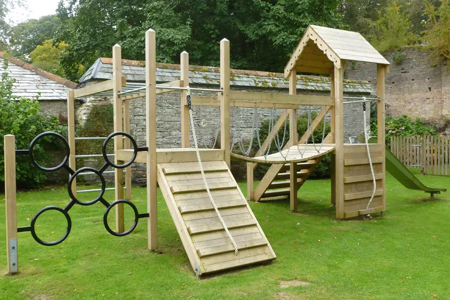 A side view of the Pencarrow Towers multi-play unit showing the wooden climbing ramp, black ring climber, chain rope net platform, stainless steel slide, and elevated tower, in a walled garden with stone walls.