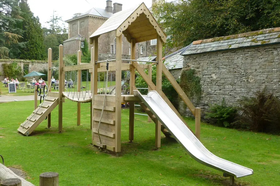 The Pencarrow Towers multi-play unit with a stainless steel slide, rope net swing, climbing ramp, and elevated tower with pitched roof, installed on a grass lawn with Pencarrow House visible in the background.