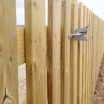 A close-up of a galvanised gate latch on a timber palisade fence with round top pickets, showing the gate post and sandy ground surface.