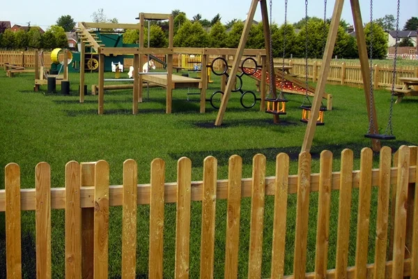 A view over a timber palisade picket fence into a well-equipped playground with a large climbing frame, swings, and other activity stations on a green artificial grass surface.
