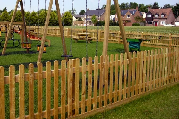 Timber palisade play area fencing with a round top picket design, enclosing a playground with a swing set and picnic bench on a green grass surface.