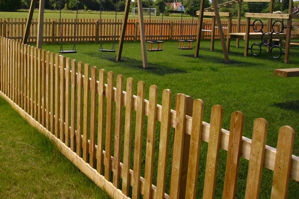A long shot of a timber palisade fence with round top pickets enclosing a large playground with swings and a climbing frame on a grassy field.