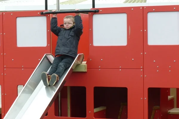 Two children on the slide of a red timber double-decker play bus in an outdoor playground.