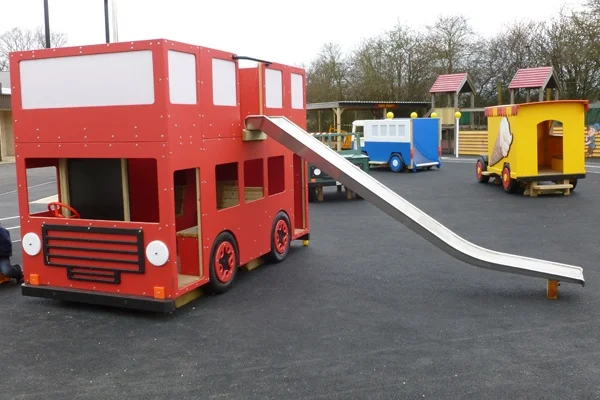 A red timber double-decker play bus with a slide, on a tarmac surface in an outdoor playground.