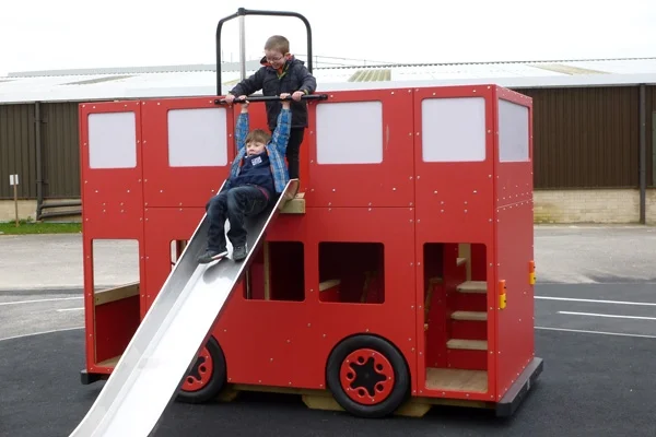 Two children on the slide of a red timber double-decker play bus in an outdoor playground.