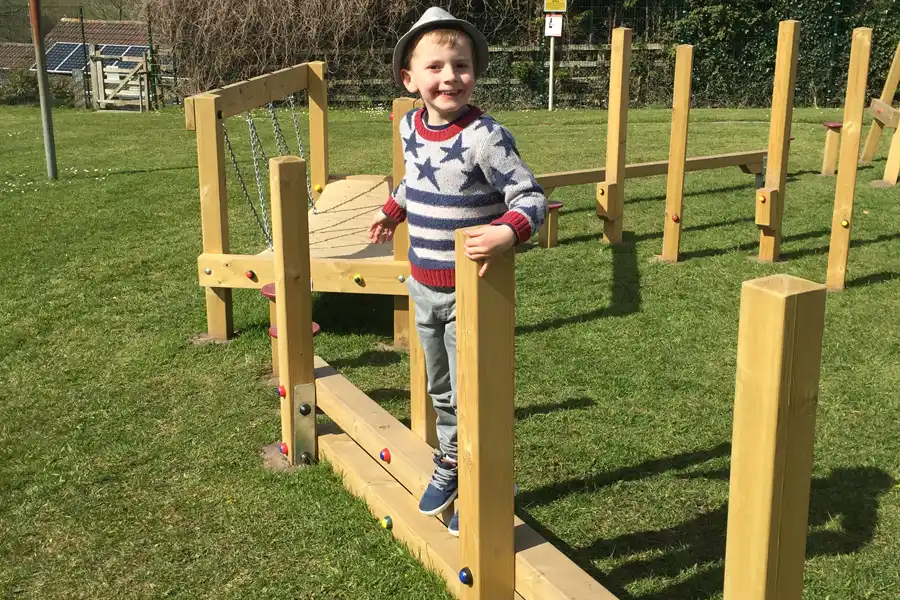 A happy young boy in a star-patterned sweater and a hat smiles while balancing on the Post Weave in an outdoor playground.