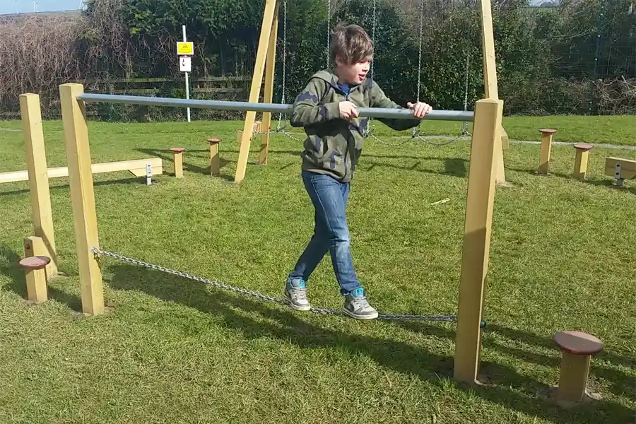 A boy carefully balances on the chain of the Postman's Bridge, holding the metal bar for support in an outdoor play park.