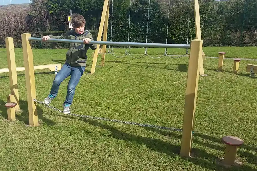 A young boy in a camouflage jacket hangs from the metal bar of the Postman's Bridge while balancing on the chain.