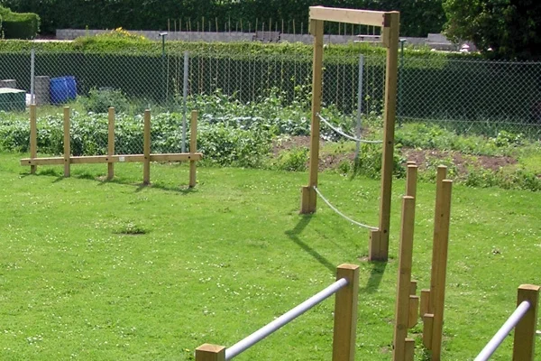 A wooden Postman's Bridge and other trim trail equipment set in a large, grassy school field with a chain-link fence in the background.