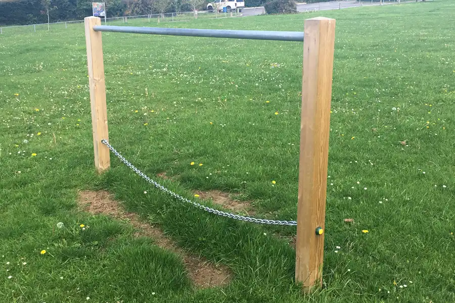 t A simple wooden frame Postman's Bridge with a metal handrail and chain, installed in a grassy field.