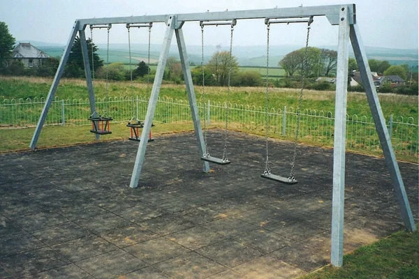 A galvanised metal quad swing frame with two cradle seats and two flat seats, set on a black rubber tile surface in a rural playground overlooking green fields.