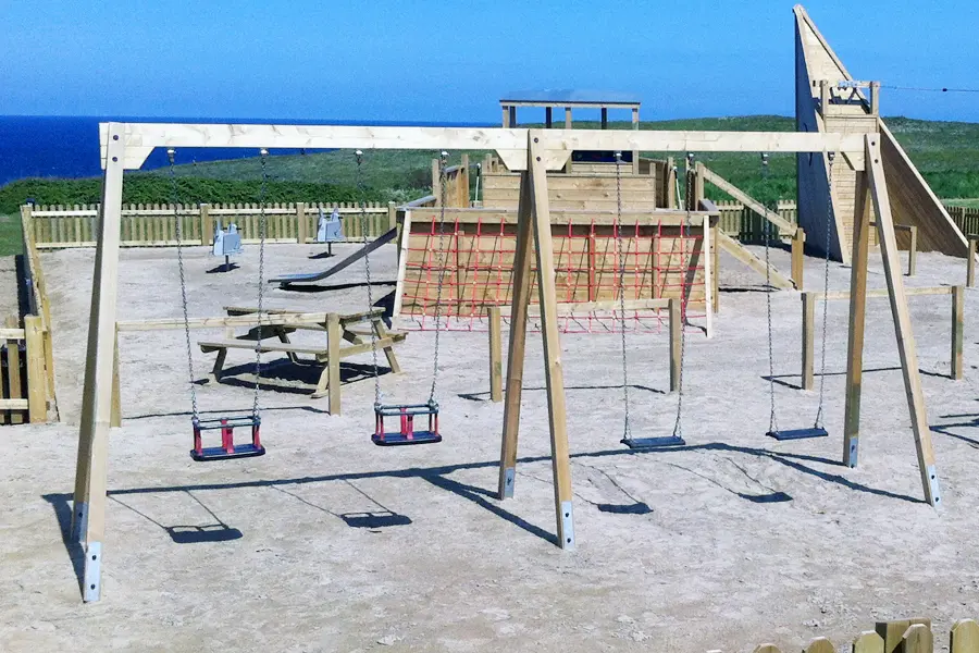 A side view of a timber quad swing frame with two flat seats and two cradle seats, located in a sandy coastal playground with a large wooden fort in the background.