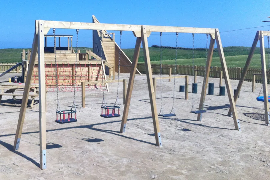 A side view of a timber quad swing frame with two flat seats and two cradle seats, located in a sandy coastal playground with a large wooden fort in the background.