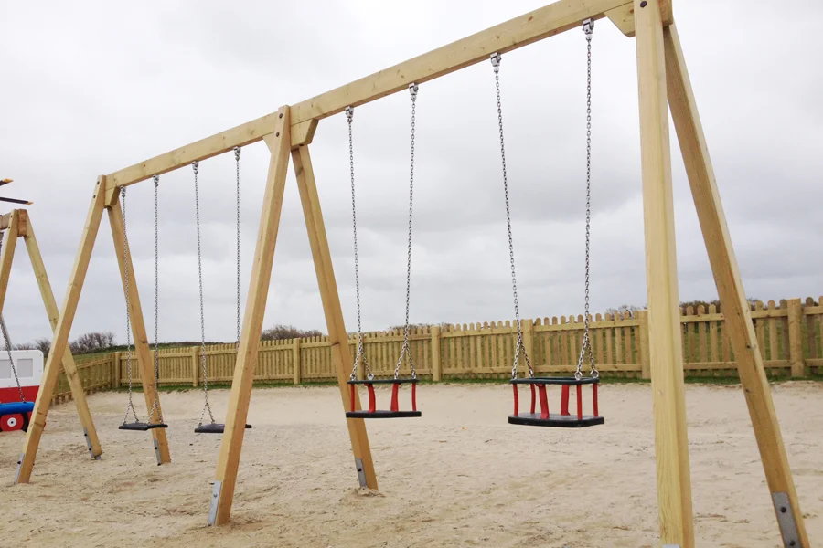 A rear-view of a timber quad swing frame with two flat seats and two cradle seats on a sandy surface, with a wooden fence and overcast sky in the background.