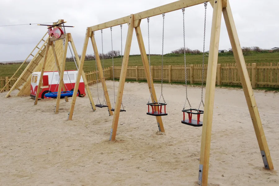 A timber quad swing with two flat seats and two cradle seats on a sandy surface, with a red and white camper van play vehicle and a zip line in the background.