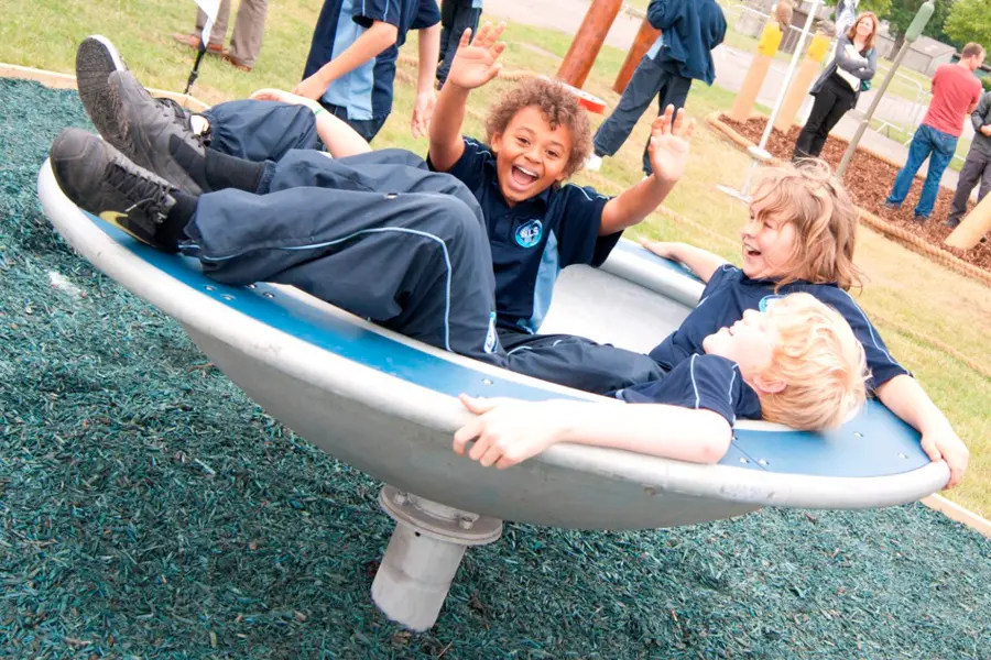 Three school children in uniform laughing and having fun while spinning in the Revolve, a multi-user bowl-shaped roundabout in a school playground.