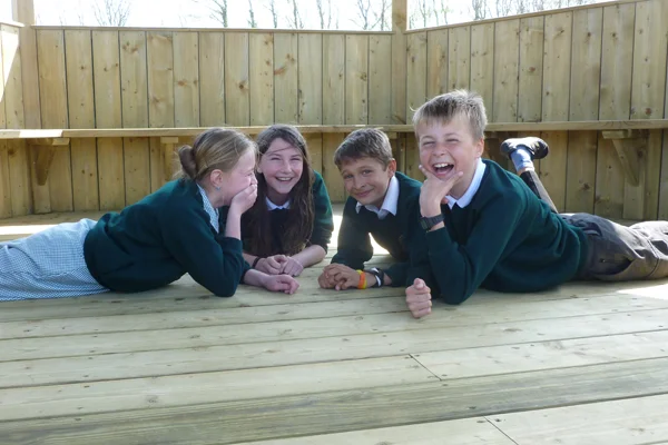 Four schoolchildren in uniform laughing and lying on the decked floor of a timber shelter stage, enjoying themselves in an outdoor classroom setting.