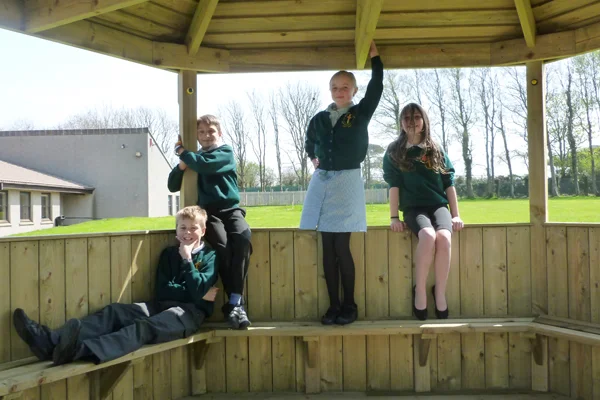 Four schoolchildren in uniform playing and posing inside a hexagonal timber shelter stage, using the benches and posts as part of their game in a school playground.