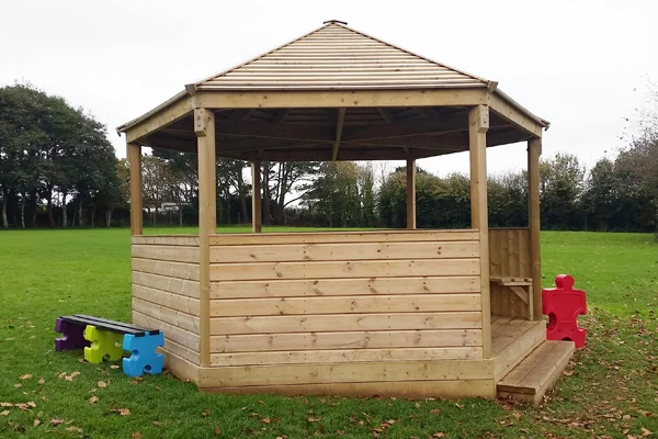 A hexagonal timber shelter and stage with a hip roof and decked floor, shown on a school playing field with colourful jigsaw-shaped seats nearby.