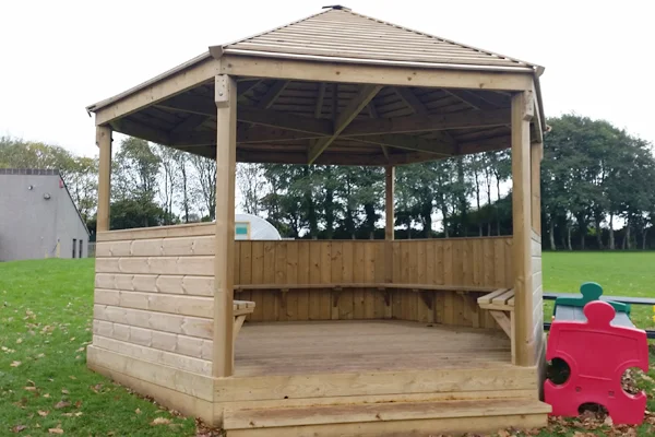 A hexagonal timber shelter and stage with a hip roof, half-height walls, and internal benches, set on a grassy school playing field, serving as an outdoor classroom.
