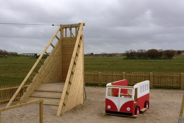 A wooden Shipwreck Cableway zip wire tower with angled ladder and zip wire cable, installed in a sandy outdoor playground next to a red and white VW campervan play unit.