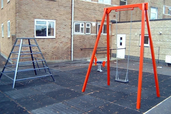 A bright orange metal A-frame single swing with a flat seat, next to a climbing frame on a black rubber tile surface in a school playground.