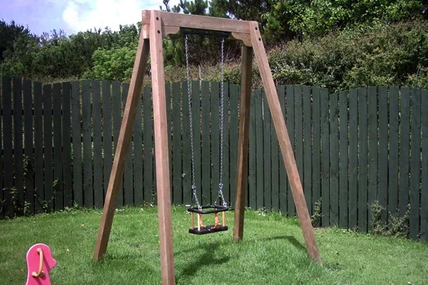 A single timber A-frame swing with a toddler seat, set in a green garden with a dark green wooden fence in the background.