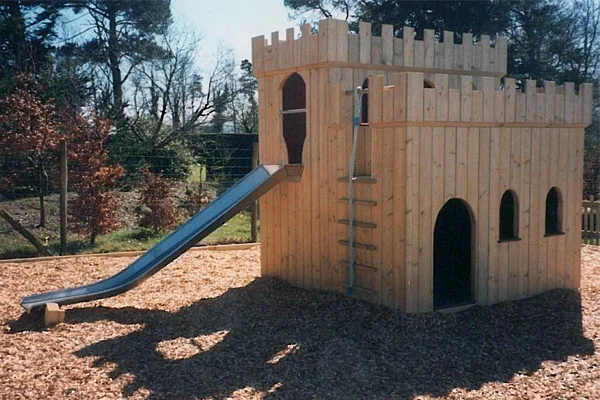 A side view of the Small Fort With Slide wooden castle outdoor play equipment, showing the stainless steel slide, rope ladder, arched doorways, and castle battlements on a wood chip playground.