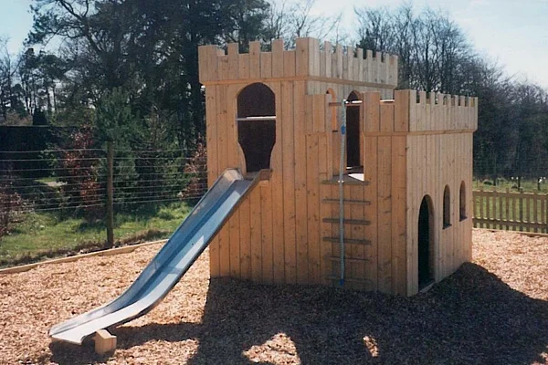 A wooden Small Fort With Slide outdoor play equipment designed as a castle with battlements, a stainless steel slide, and a rope ladder, installed on a wood chip playground surface.