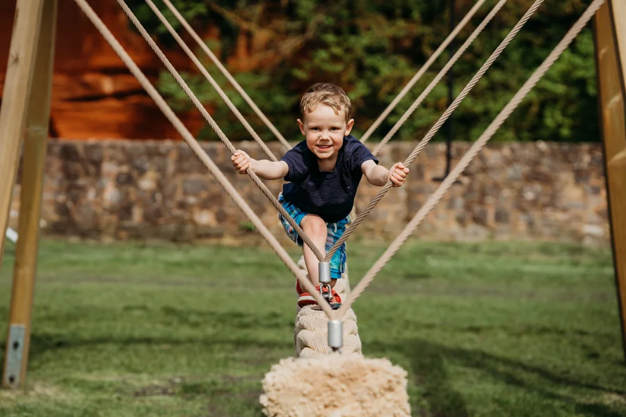 A young boy smiling as he balances on a thick, braided snake rope swing, holding onto the support ropes in a grassy outdoor playground.